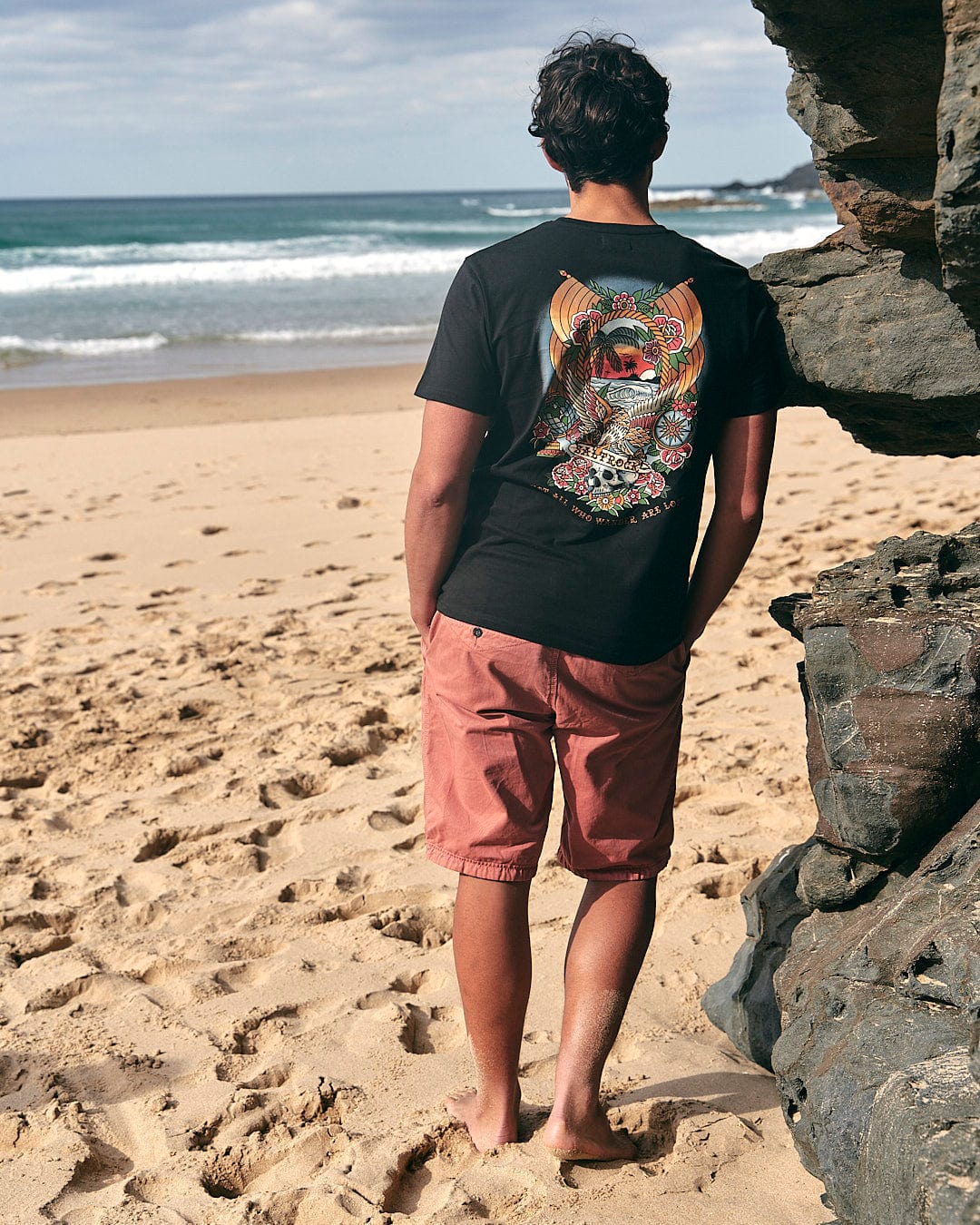 A man standing on the beach wearing a Saltrock Tattoo Island Mens Short Sleeve T-Shirt in Black.