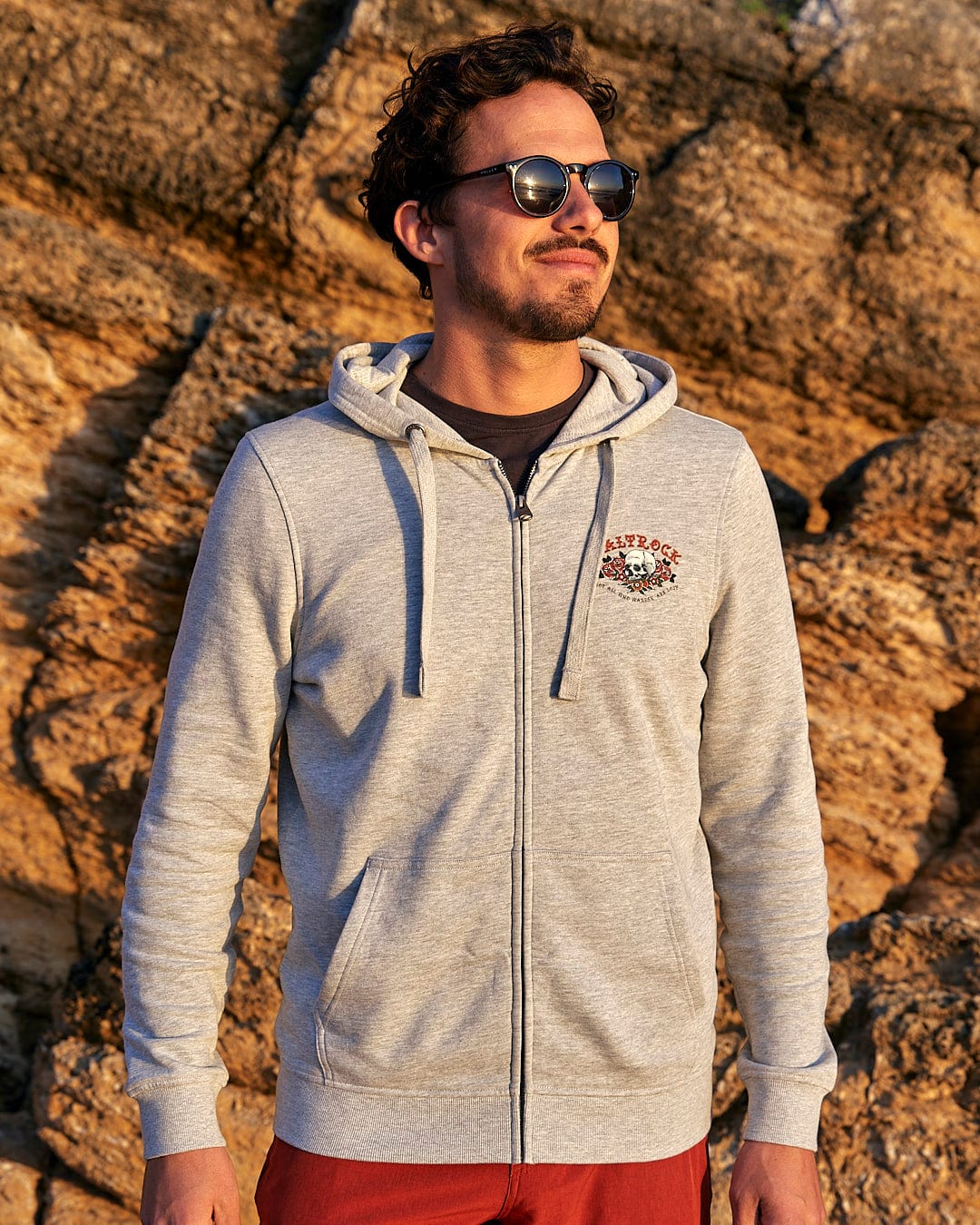 A man wearing a Saltrock Tattoo Island - Mens Zip Hoodie - Grey standing in front of rocks.
