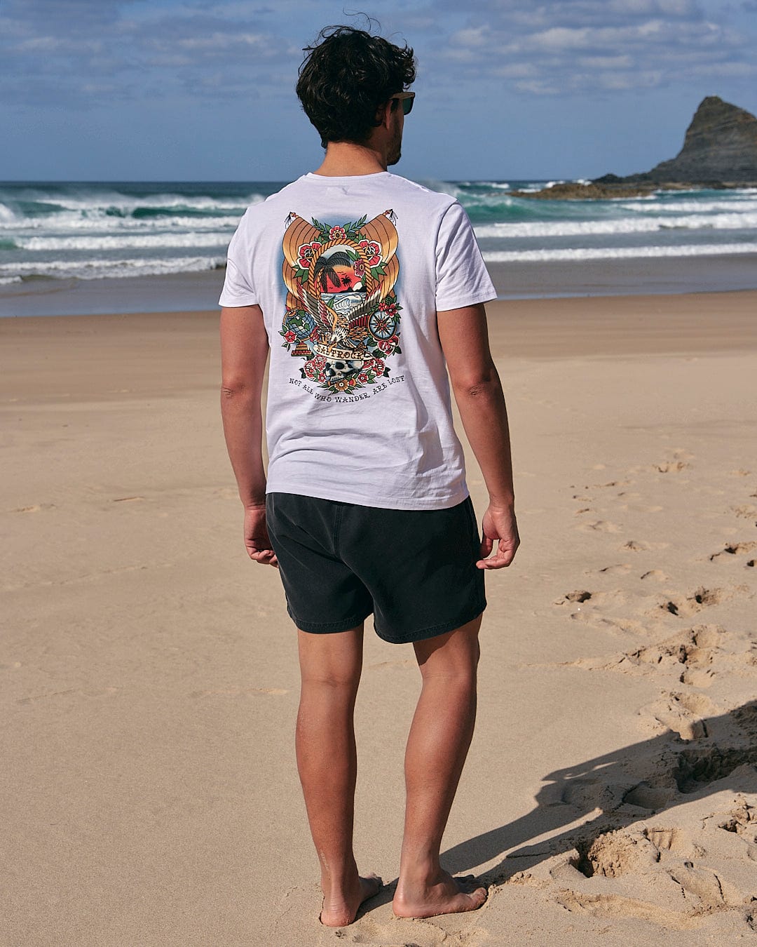 A man standing on the beach wearing a Saltrock - Tattoo Island Mens Short Sleeve T-Shirt in White and shorts.