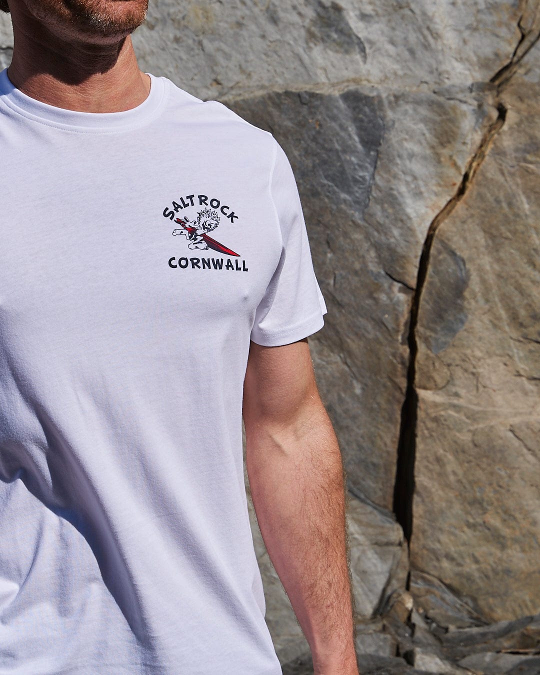 A man in a Wave Rider Cornwall - Mens Short Sleeve T-Shirt in White featuring Saltrock branding stands against a rocky backdrop.