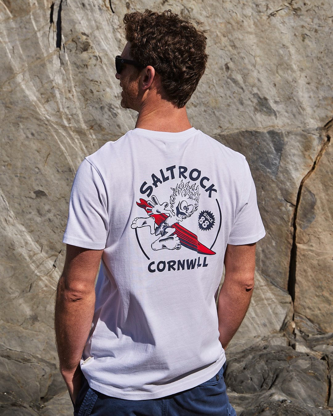A man wearing sunglasses and a Wave Rider Cornwall Mens Short Sleeve T-shirt in white made of peached soft material with a Saltrock logo stands facing a rocky background.