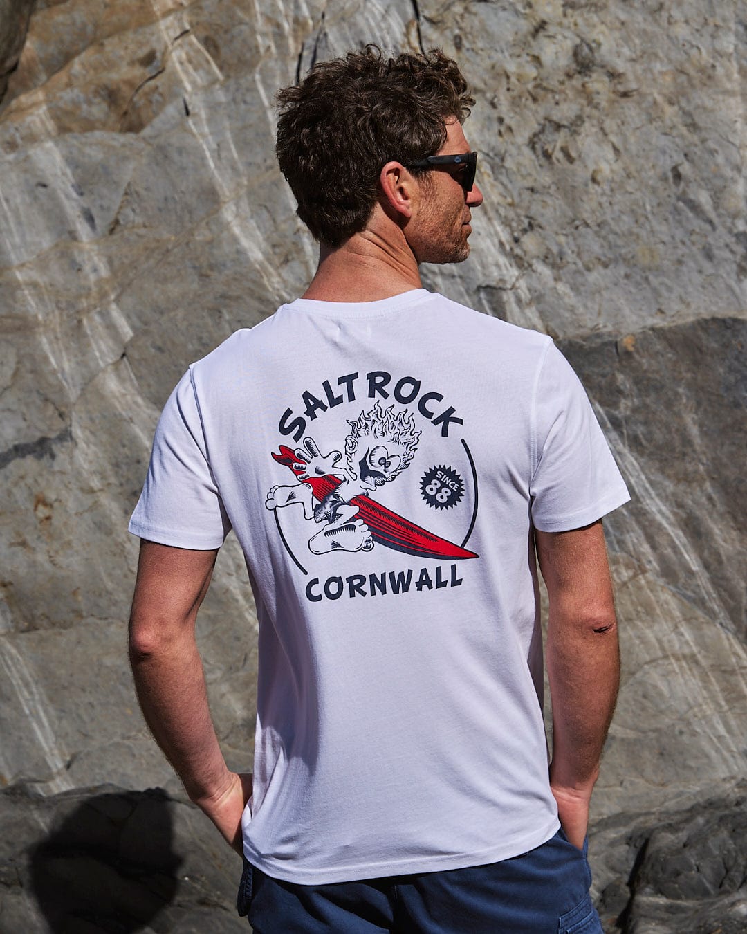 Man in sunglasses standing against a rocky backdrop wearing a white t-shirt with "Wave Rider Cornwall" graphic design, crafted from peached soft material by Saltrock.