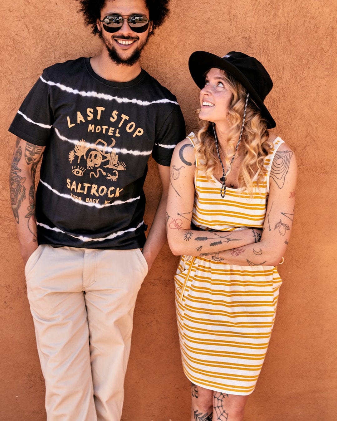 A smiling man and a woman wearing a Saltrock Skylar Bauhaus - Womens Stripe Dress in yellow and casual summer clothes stand against an orange wall, looking at each other.