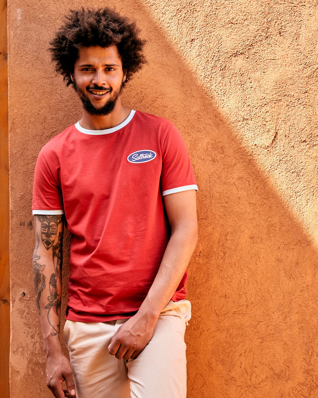 A young man with curly hair and a tattoo on his arm smiles while leaning against an orange wall, dressed in a red Striker Ringer - Mens Short Sleeve T-Shirt with Saltrock branding embroidered and beige pants.