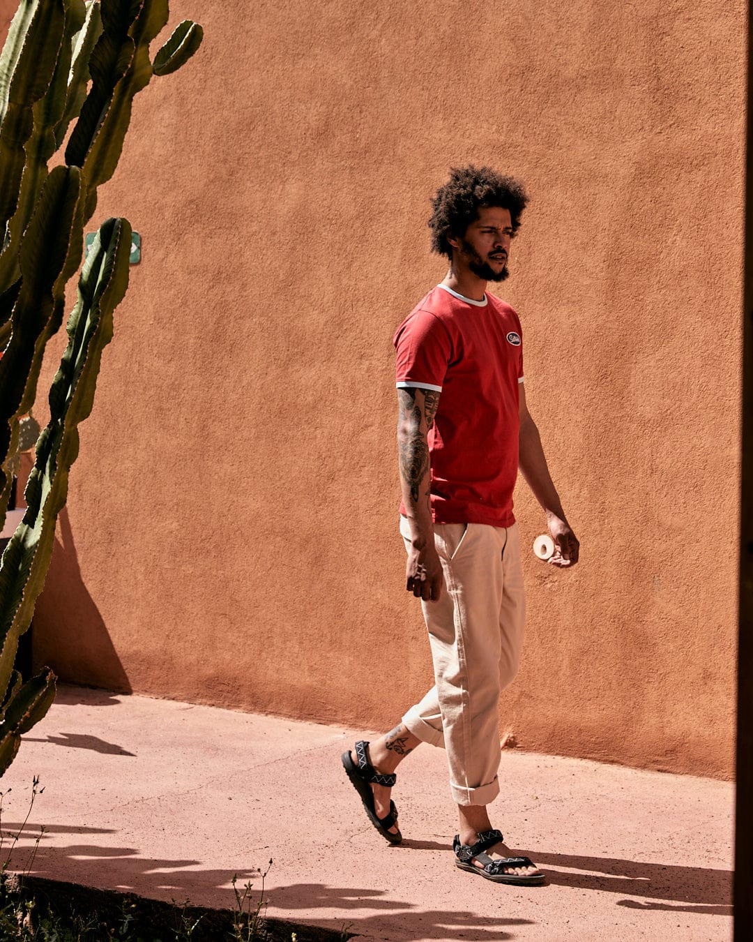 A man with curly hair walking past a cactus, wearing a 100% cotton red Striker Ringer t-shirt with Saltrock embroidered branding, beige trousers, and sandals against an earthy wall.
