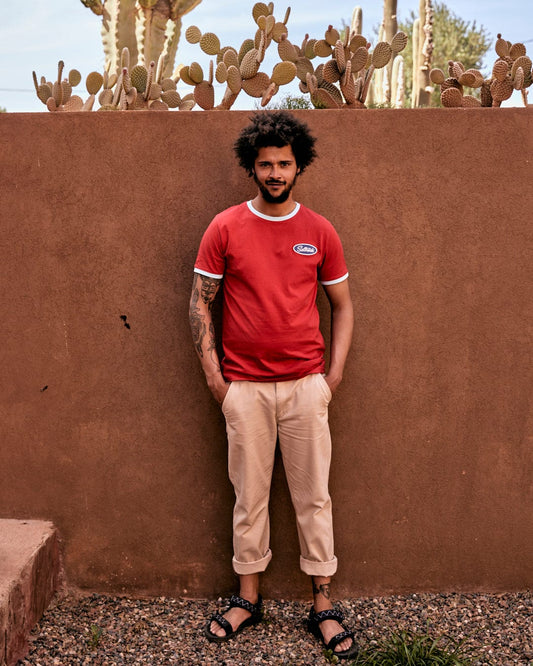 A man with curly hair and a tattoo on his arm stands in front of a brown wall, wearing a red Striker Ringer - Mens Short Sleeve T-Shirt with Saltrock embroidered branding and beige pants, with cacti in the background.