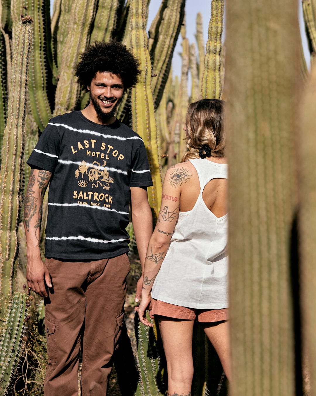 A man and a woman with tattoos walking among tall cacti in a desert setting. The man smiles at the camera, both dressed in casual summer clothes featuring Saltrock's Last Stop Motel - Mens Short Sleeve Tie Dye T-Shirt in Dark Grey print.