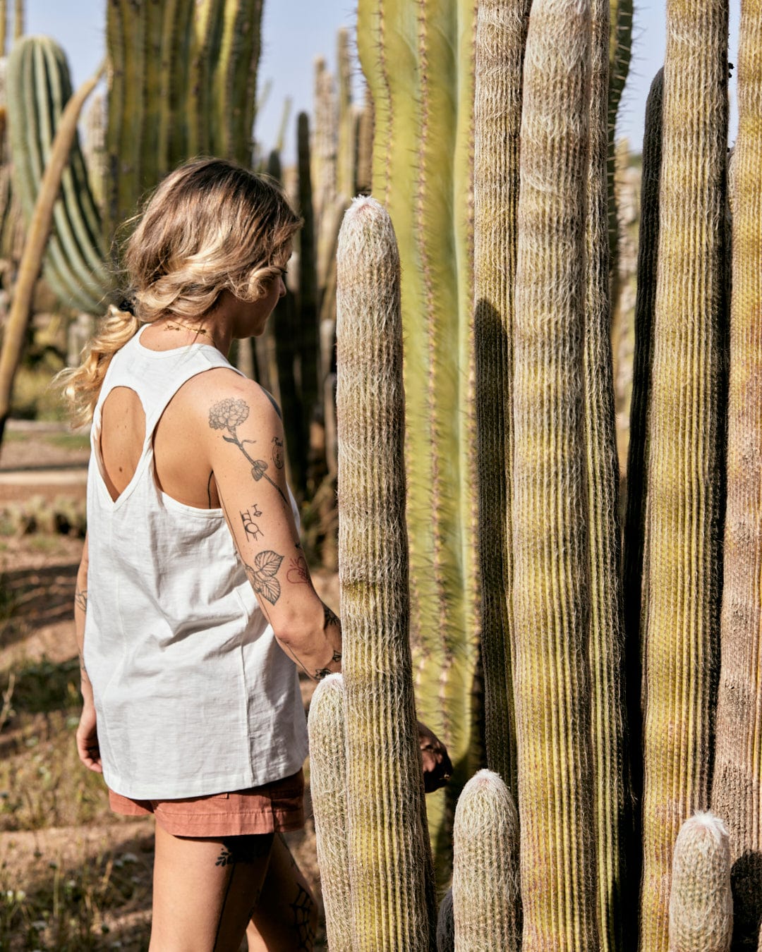 A woman with a tattoo on her arm examines tall cacti in a sunny desert landscape. She wears a Saltrock Palmera Womens Vest in White with a crew neckline and pink shorts.