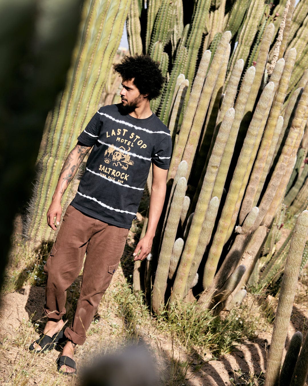 Man with an afro wearing a Last Stop Motel - Mens Short Sleeve Tie Dye T-Shirt in Dark Grey with crew neckline and brown pants standing near tall cacti in a sunny outdoor setting. (Brand: Saltrock)