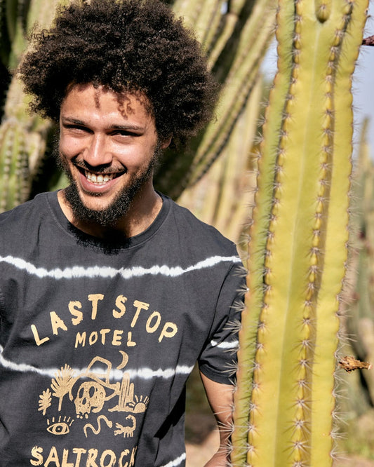 Young man with curly hair smiling, wearing a Last Stop Motel - Mens Short Sleeve Tie Dye T-Shirt in Dark Grey with Saltrock branding, standing near tall cacti in a sunny outdoor setting.