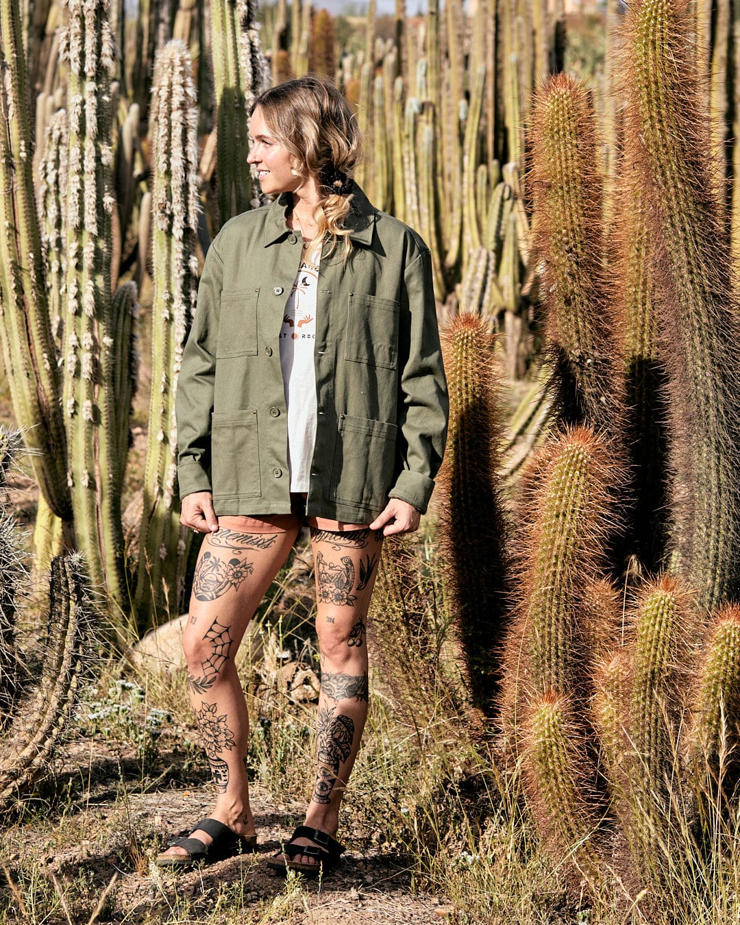 Woman with tattoos standing among tall cacti in a desert landscape, wearing a Saltrock Barden - Womens Lightweight Utility Jacket in Dark Green and shorts.