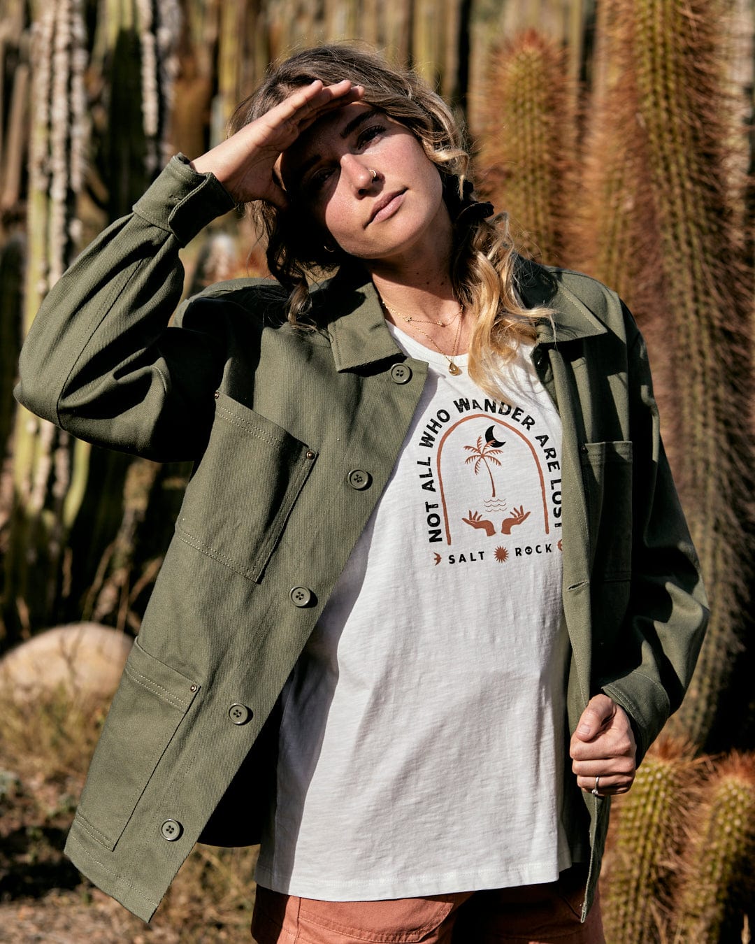 A woman in a forest, wearing a Saltrock Barden - Womens Lightweight Utility Jacket in Dark Green and white t-shirt with text, shielding her eyes from the sun.