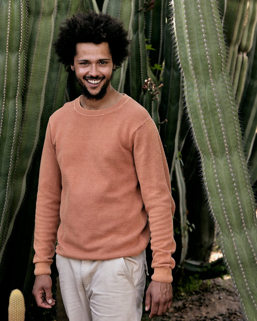 A smiling man with an afro wearing a Saltrock Moss - Mens Washed Knitted Crew in Orange stands in front of tall cactus plants.