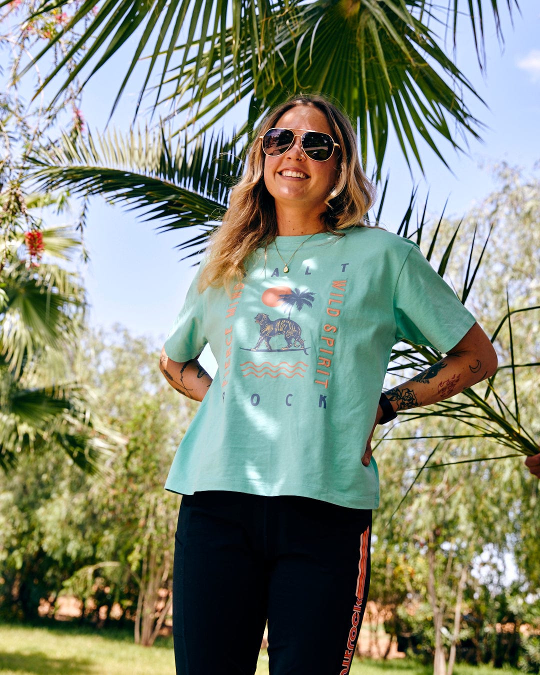 A woman stands smiling under palm trees, wearing a teal Saltrock Fierce Mind - Womens Boxy T-Shirt - Green, sunglasses, and black trousers, with her hands in her pockets.