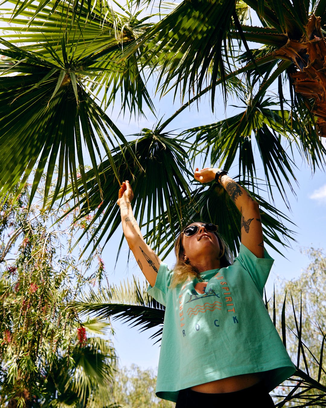 Woman reaching for a fruit on a palm tree, wearing sunglasses and a Saltrock Fierce Mind - Womens Boxy T-Shirt in Green with wild spirit graphics, under a blue sky.