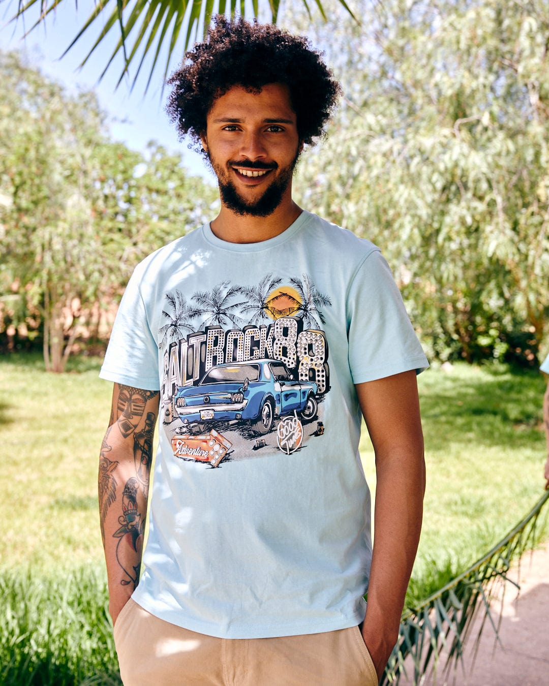 A man with curly hair, wearing a Saltrock Neon Boneyard t-shirt and khaki shorts, smiles while standing outdoors in a sunny, garden-like setting.