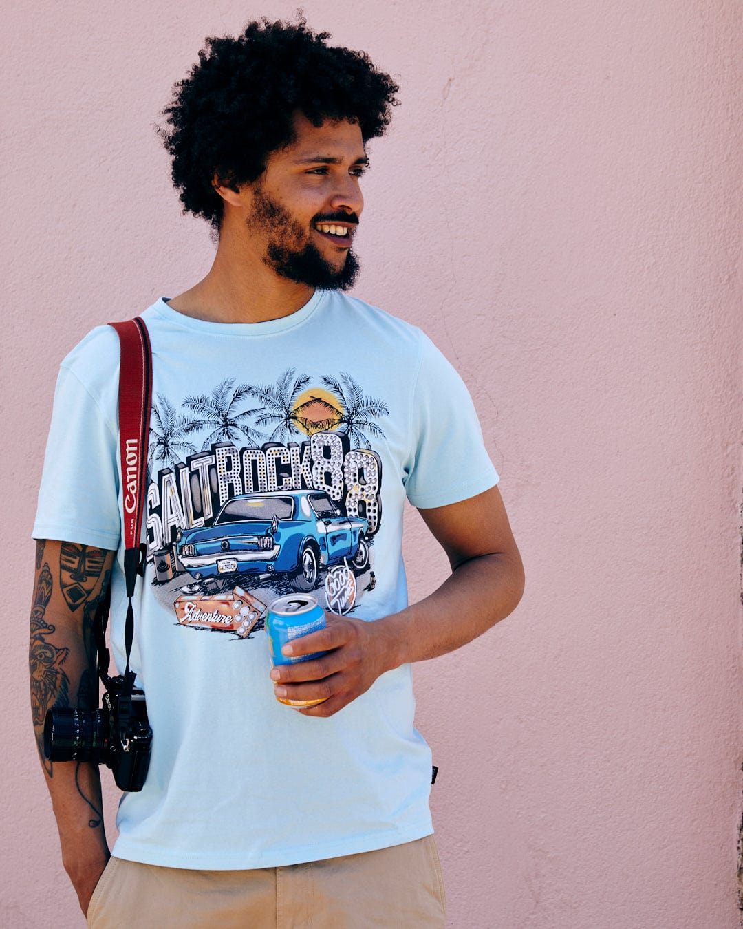 A man with curly hair, holding a beverage can, stands against a pink wall wearing a Saltrock Neon Boneyard - Mens Short Sleeve T-Shirt in Light Blue and carrying a red shoulder bag.