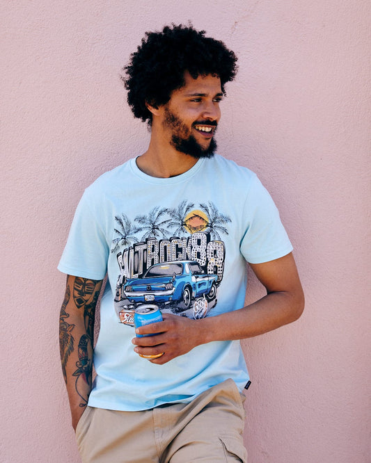 A man leaning against a pink wall, holding a drink, wearing a Neon Boneyard - Mens Short Sleeve T-Shirt in Light Blue from Saltrock and beige shorts, smiling slightly.