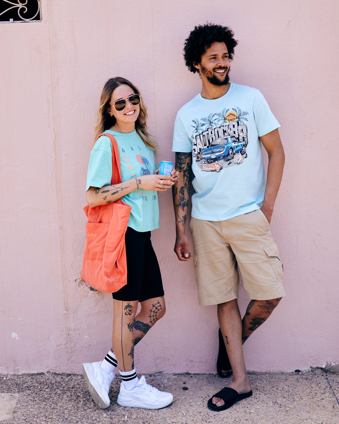 A man and woman smiling and standing against a pink wall, the woman holding a drink and a Neon Boneyard - Mens Short Sleeve T-Shirt - Light Blue tote bag made of 100% Cotton from Saltrock, and the man with a cup and phone. Both are casually.