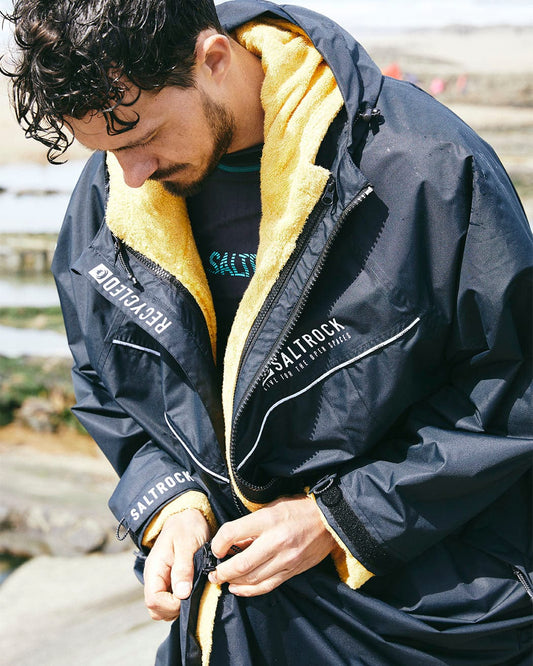 A man standing outdoors adjusts the zipper on his black and yellow Saltrock 3 in 1 Recycled Changing Robe, made from recycled material, looking down at it.