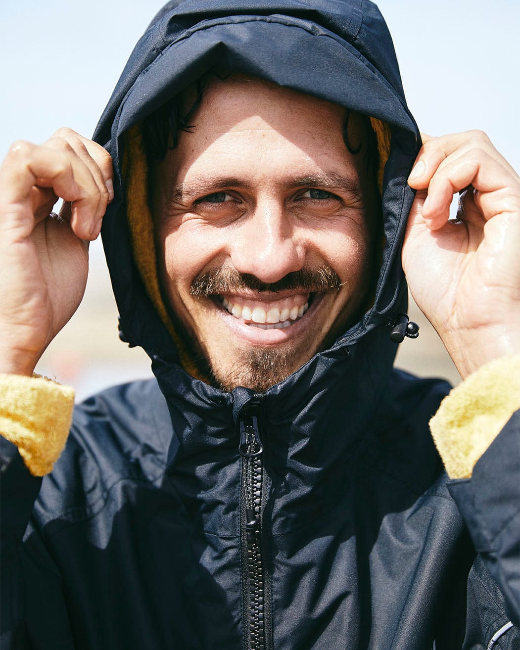 A man with a beard smiling brightly, holding the hood of his Saltrock 3 in 1 Recycled Four Seasons Changing Robe - Black/Yellow around his face.