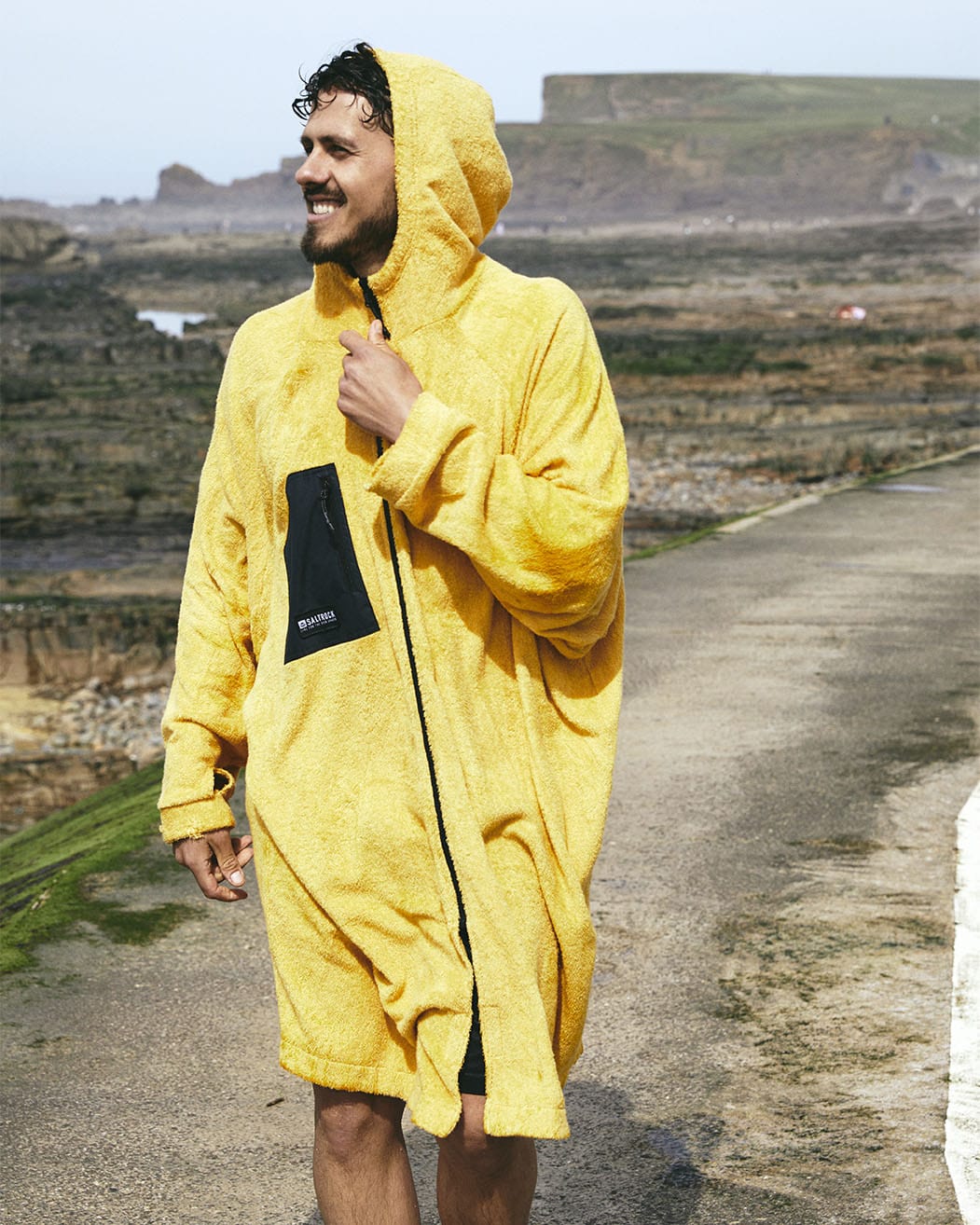 A man in a Saltrock 3 in 1 Recycled Four Seasons Changing Robe - Black/Yellow smiles while standing on a coastal path, cliffs and the ocean in the background.