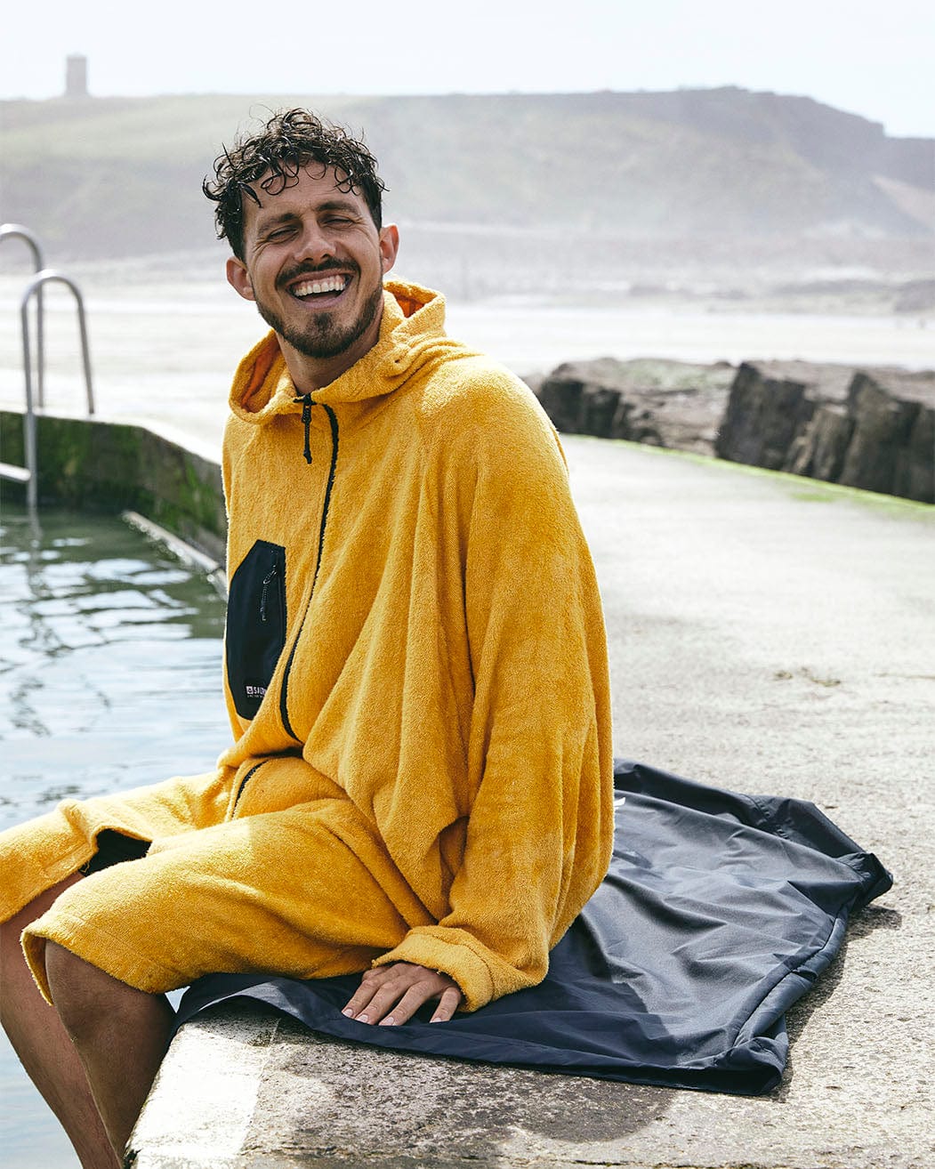 A man smiling while sitting on a pier, wearing a Saltrock 3 in 1 Recycled Four Seasons Changing Robe in Black/Yellow, with a rugged coastline and a distant lighthouse in the background.