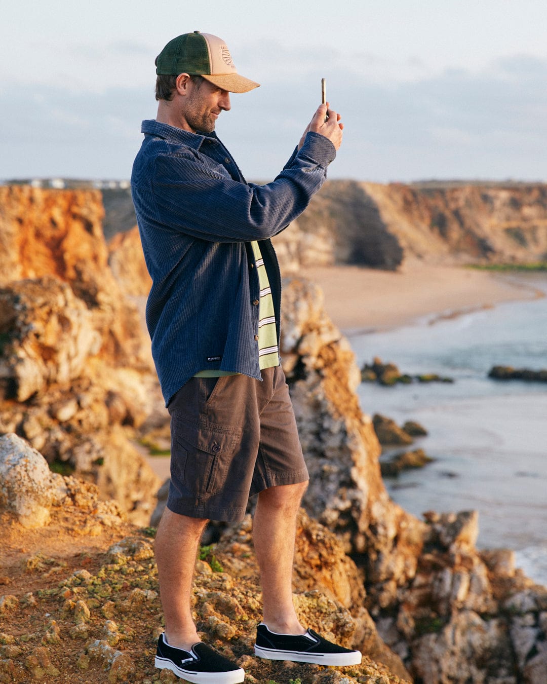A man wearing a Saltrock Ace - Mens Long Sleeve Shirt in Blue with front chest pockets standing on a cliff taking a picture of the ocean.
