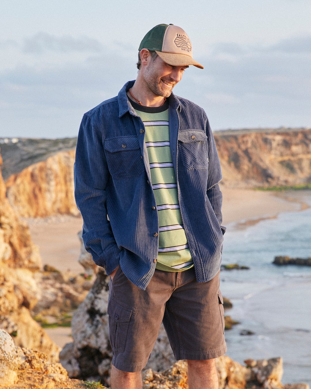 A man standing on a rocky beach wearing a Saltrock Ace - Mens Long Sleeve Shirt in Blue, hat, and shorts.