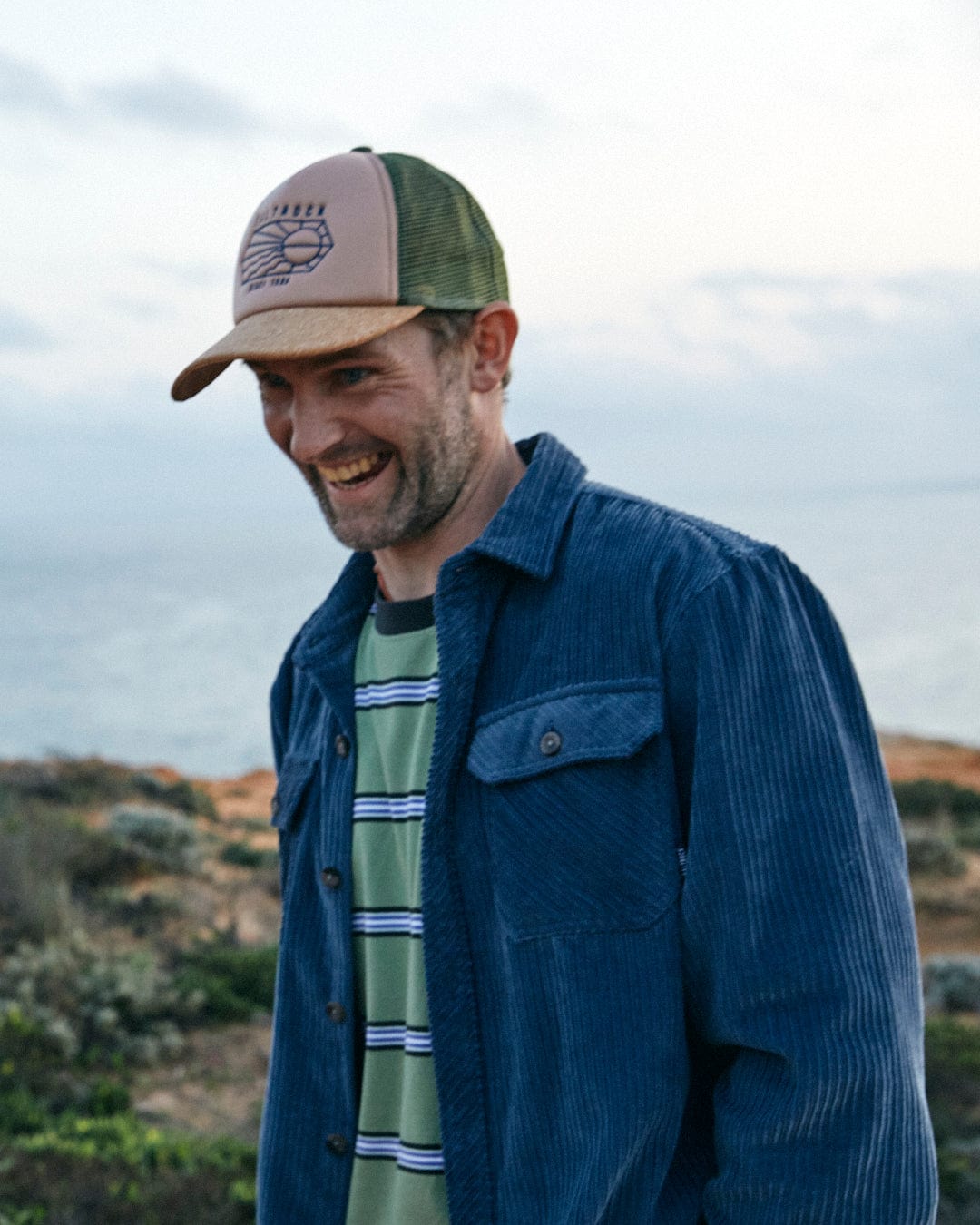 A man in a Lineal Trucker Cap in Cream with Saltrock branding and denim jacket smiling outdoors with a natural landscape in the background.