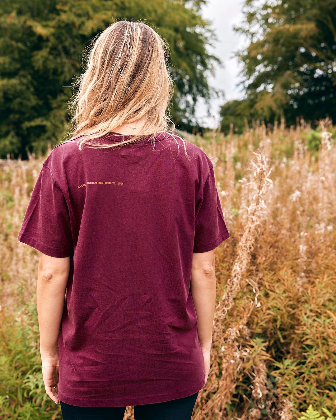 A woman wearing an Adventure Awaits - Womens Short Sleeve T-Shirt - Dark Purple by Saltrock standing in a field.