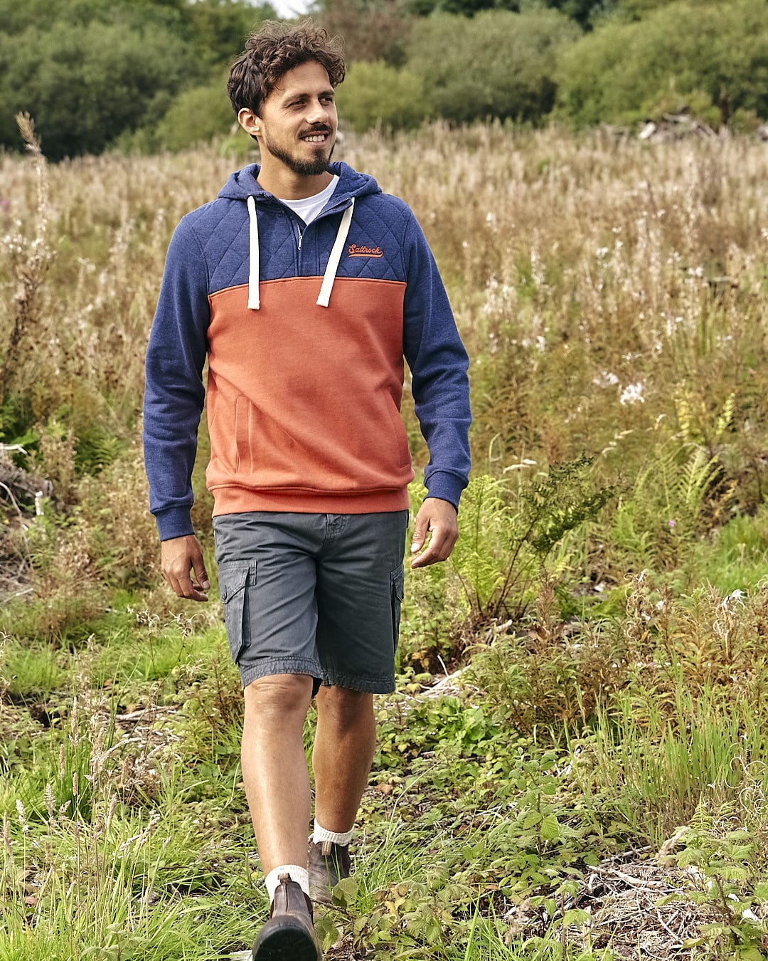 A man walking through a field wearing a Saltrock - Aiken - Mens 1/4 Neck Hoodie - Orange and shorts.