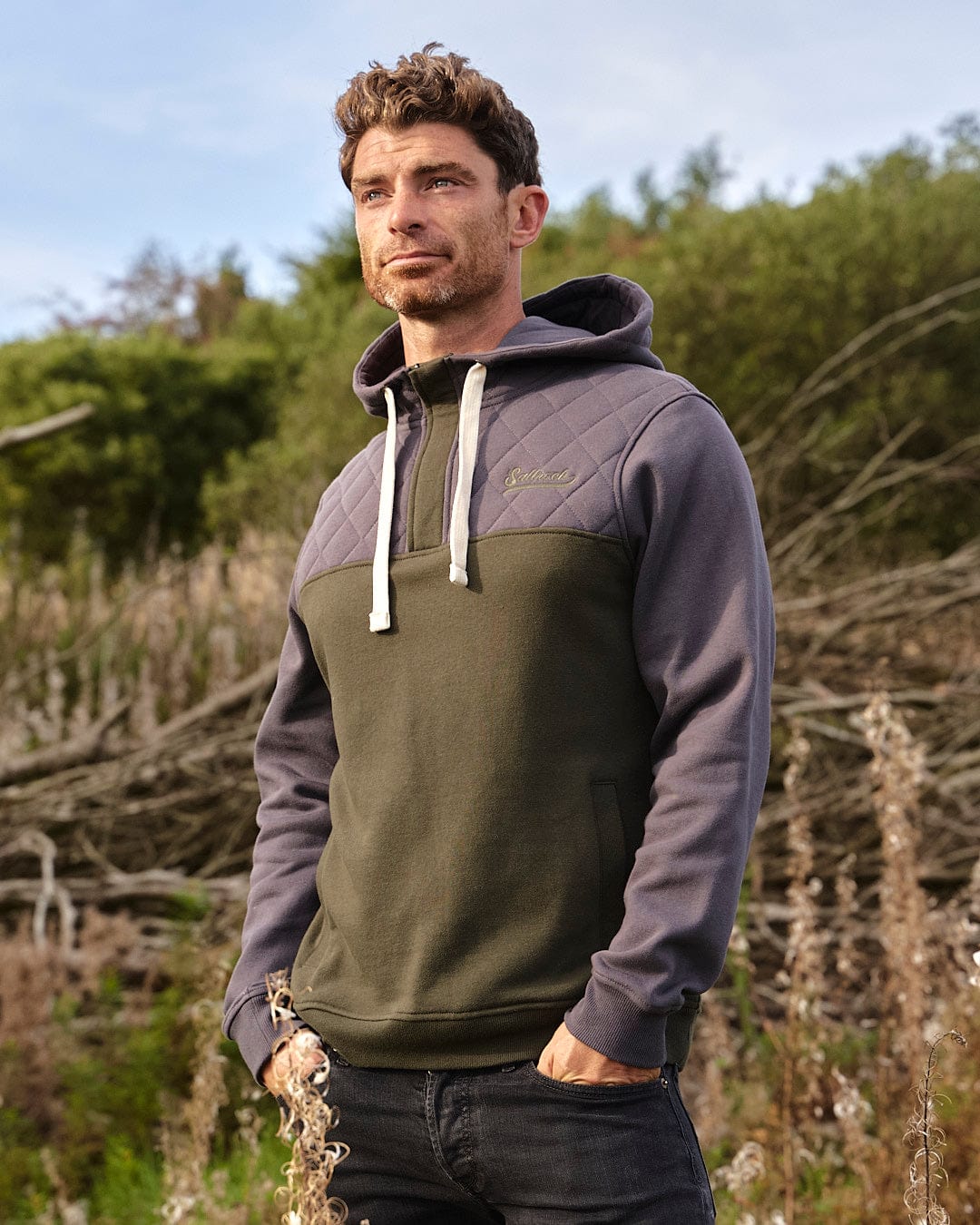 A man standing outdoors in a field, finding warmth and comfort with the Saltrock Aiken Mens 1/4 Neck Hoodie - Green.