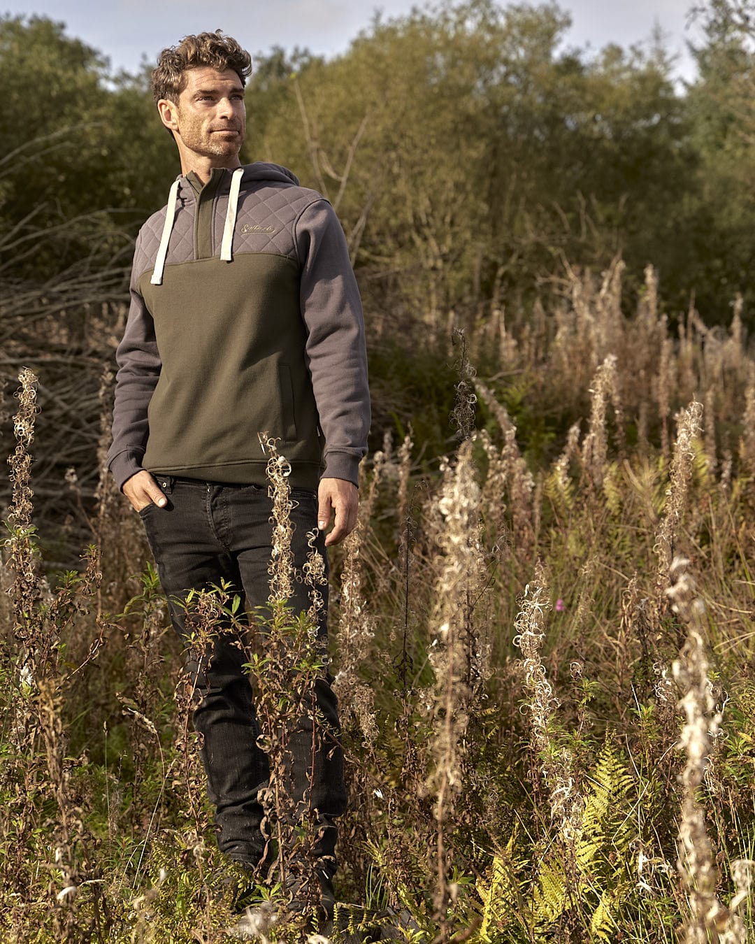 A man standing in a field, enjoying the outdoors with tall grasses while feeling warmth and comfort in his Saltrock Aiken Mens 1/4 Neck Hoodie - Green.