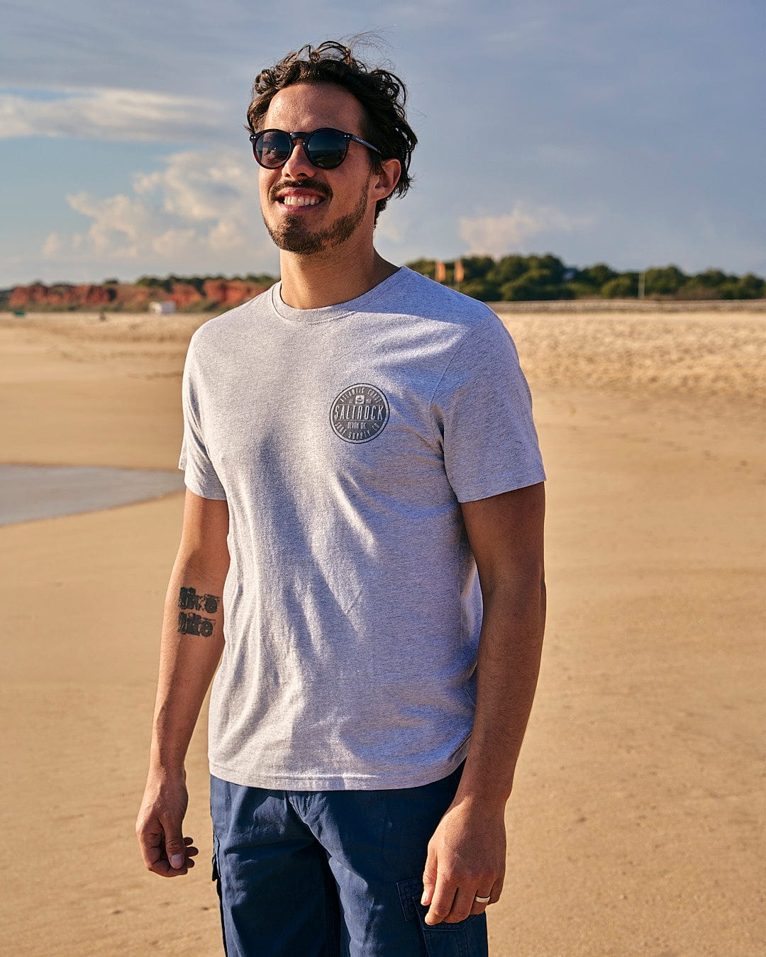 A man standing on a beach wearing Saltrock sunglasses and an All Terrain - Mens Short Sleeve T-Shirt - Grey.