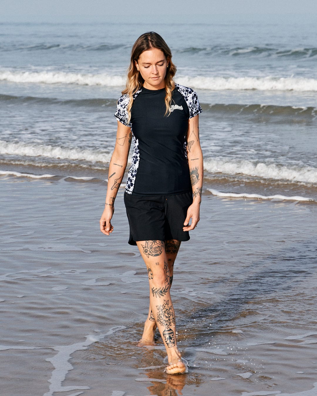 A woman with tattoos walks along the shoreline, wearing a Saltrock Amphibian Womens Boardshort in Black, focused on the water at her feet.