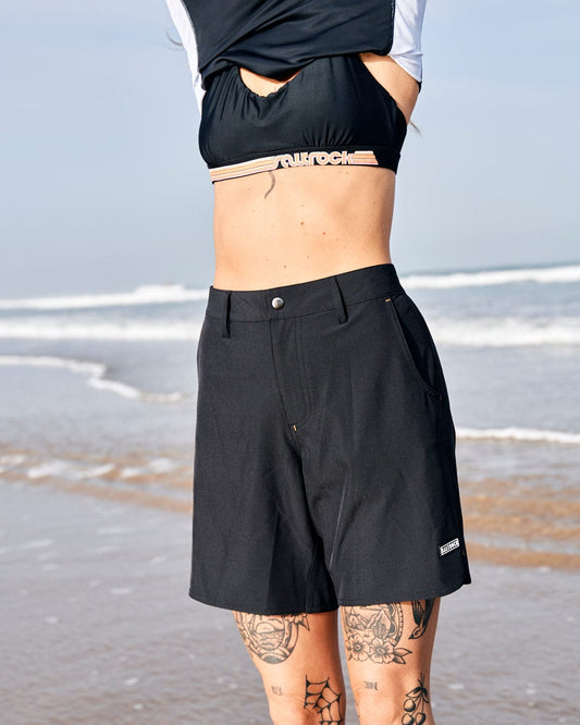 Woman on the beach wearing Saltrock's Amphibian - Womens Boardshort - Black with belt loops and a cropped top, revealing a tattooed lower back. The ocean and sky are visible in the background.
