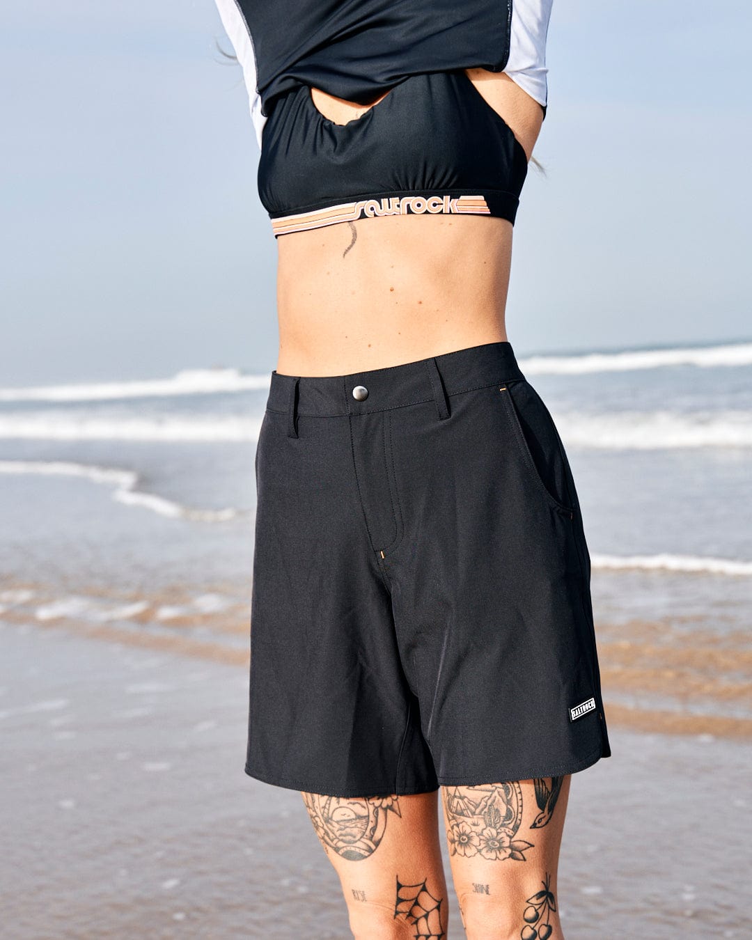 Woman on the beach wearing Saltrock's Amphibian - Womens Boardshort - Black with belt loops and a cropped top, revealing a tattooed lower back. The ocean and sky are visible in the background.
