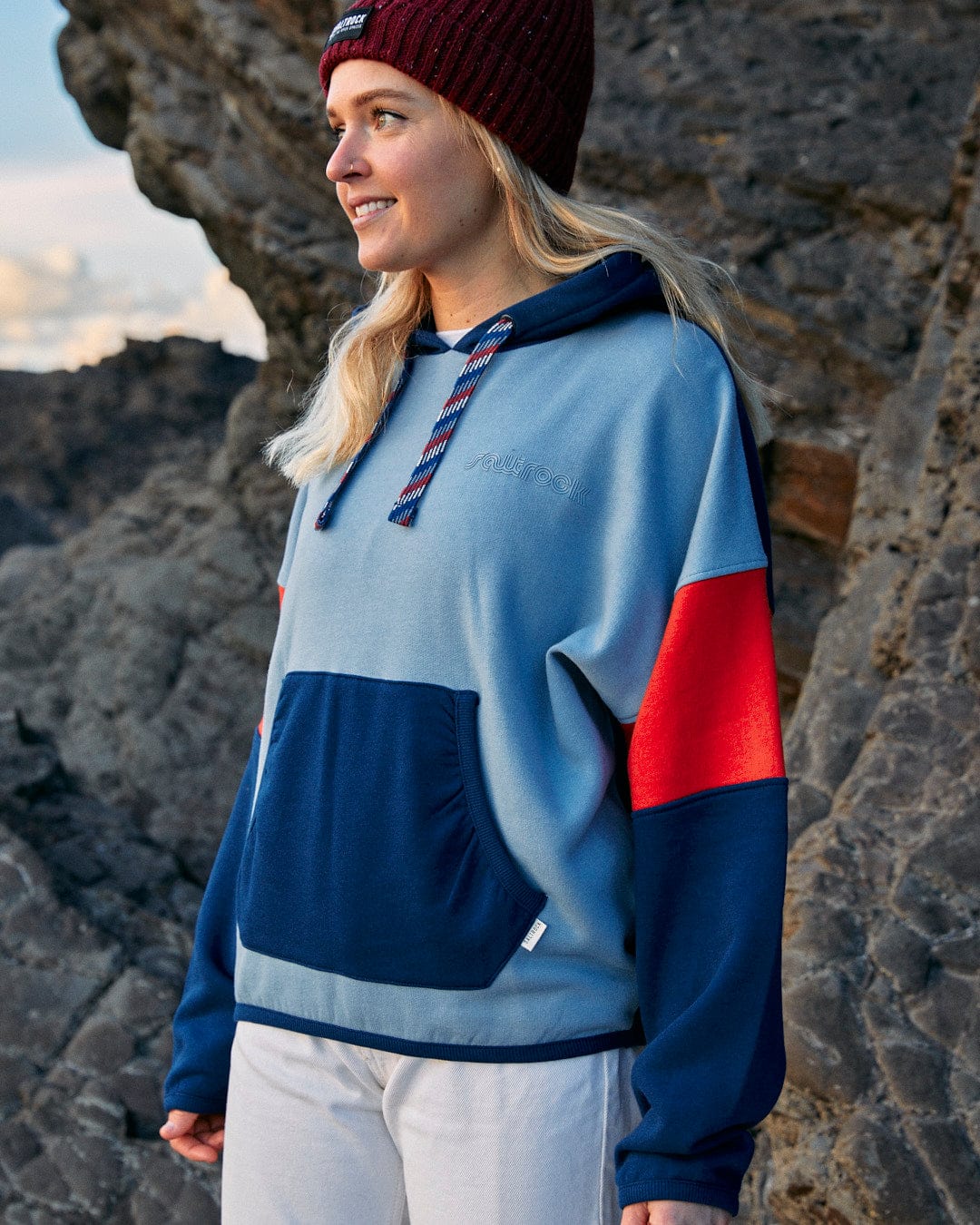 A woman wearing the Anya - Womens Pop Hoodie - Blue with Saltrock branding, standing on a rock.