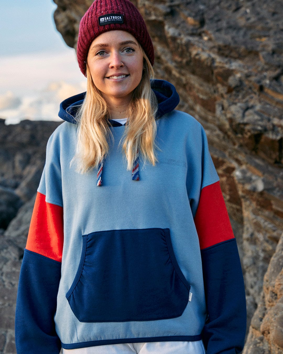 A woman wearing a relaxed fit blue and red hoodie with contrast paneling, showcasing Saltrock branding, standing on a rock.