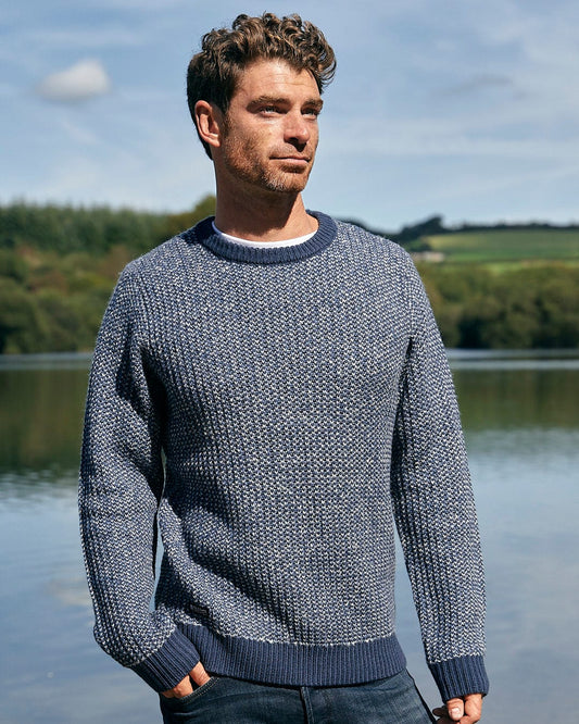 A man is standing next to a lake wearing a Saltrock Arlen Mens Crew Knit Sweater in Blue.