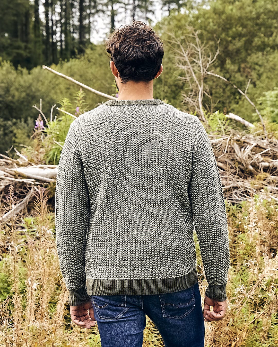 The back of a man standing in a wooded area wearing the Saltrock Arlen - Mens Crew Knit - Dark Green sweater.