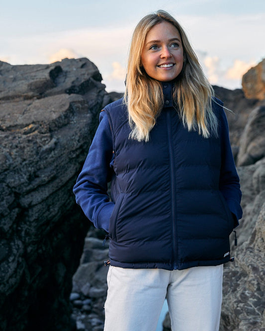 A woman standing on a rocky beach wearing a Saltrock Astra - Womens Reversible Padded Gilet - Blue/Pink.