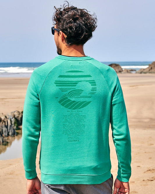 A man is standing on the beach wearing a Saltrock - Atlantic Mens Crew Sweat - Dark Green sweatshirt.