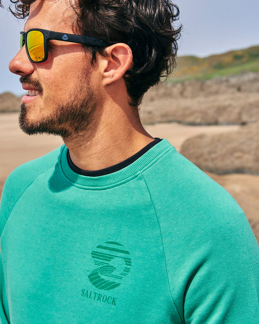 A man wearing sunglasses and a Saltrock Atlantic - Mens Crew Sweat - Dark Green on the beach.