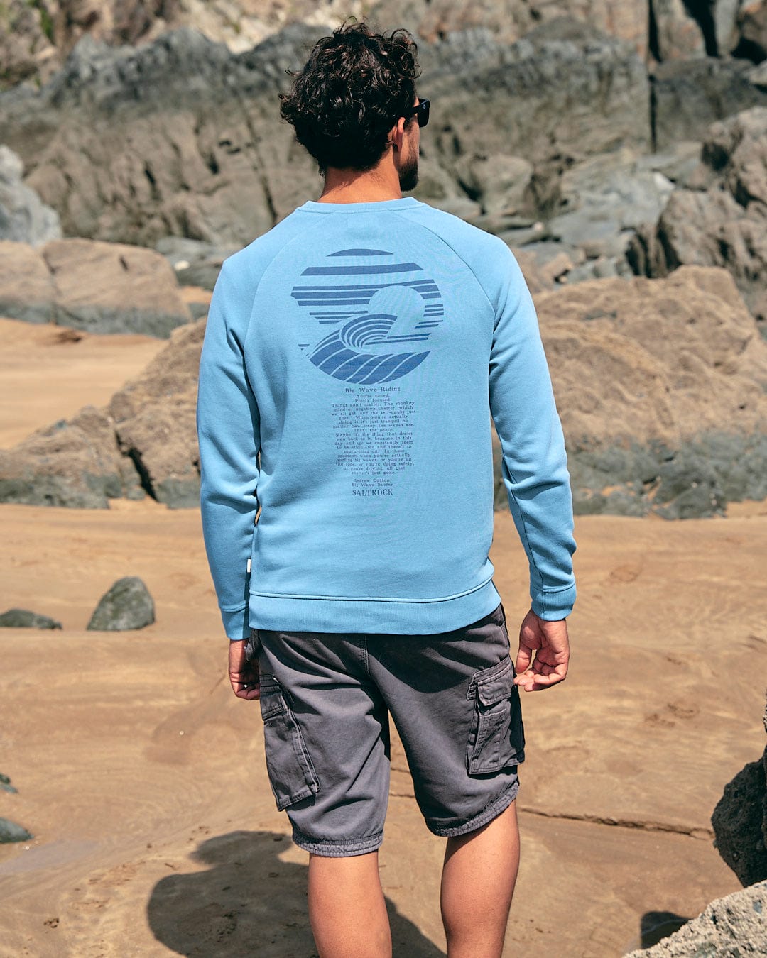 A man standing on the beach wearing a Saltrock - Atlantic Mens Crew Sweat - Light Blue.