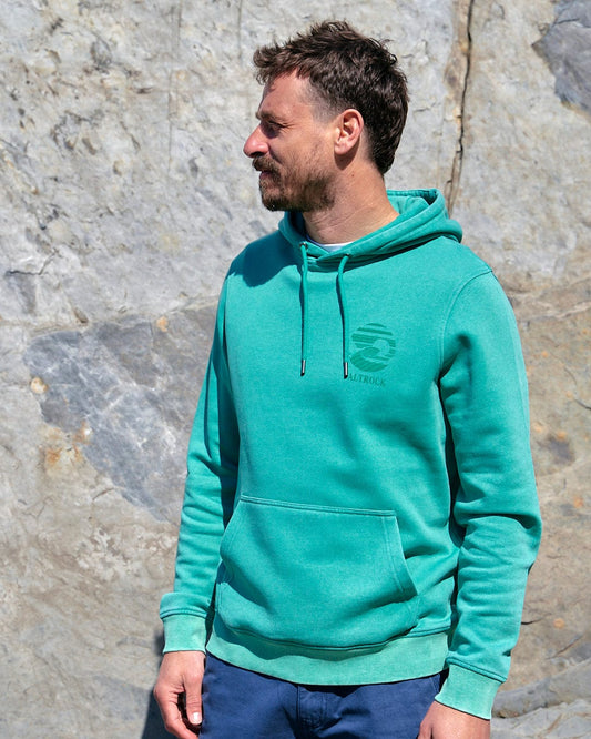 A man wearing a Saltrock Atlantic - Mens Pop Hoodie - Dark Green standing in front of a stone wall.