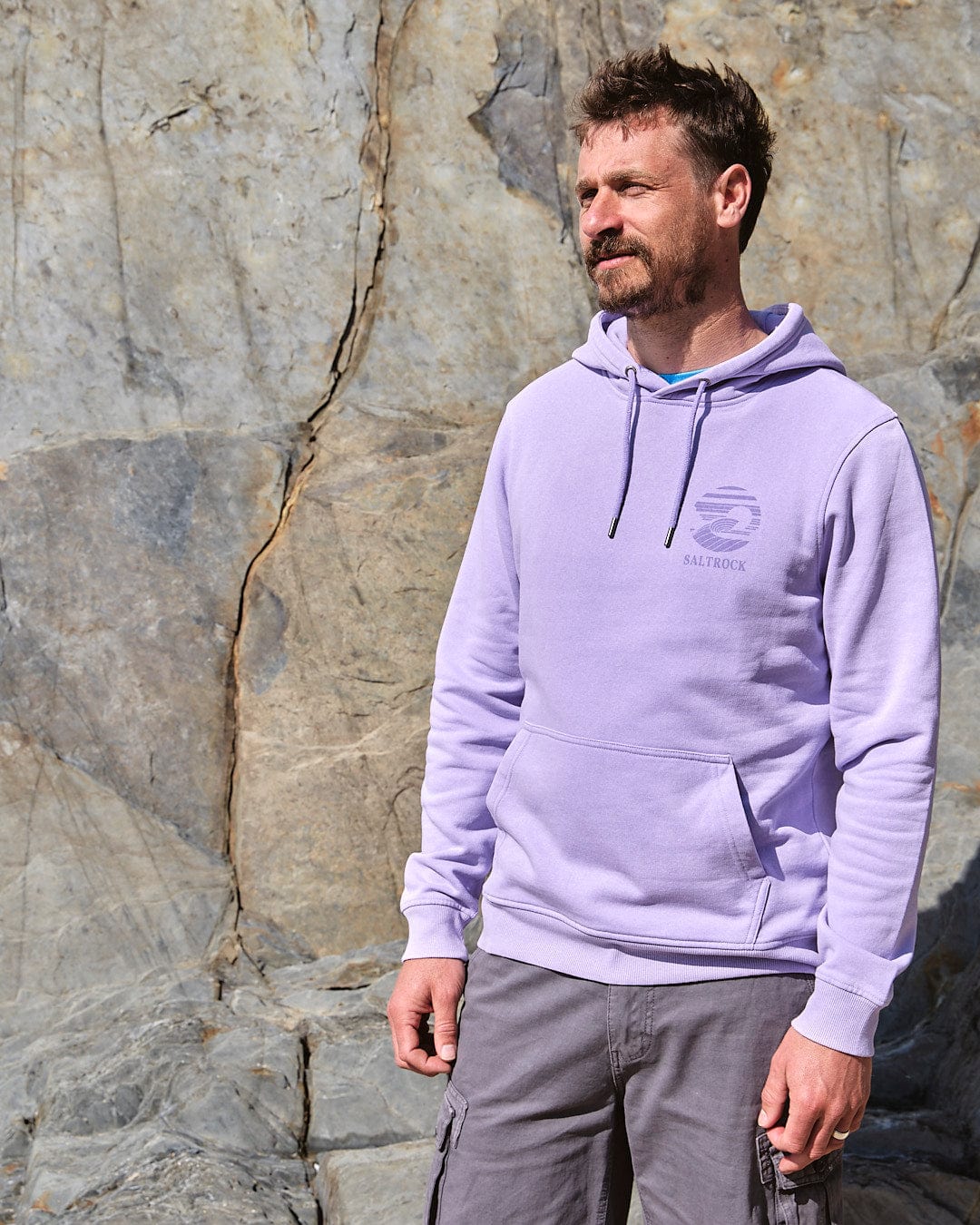 A man wearing a Saltrock Atlantic - Mens Pop Hoodie - Purple standing next to a rock wall.