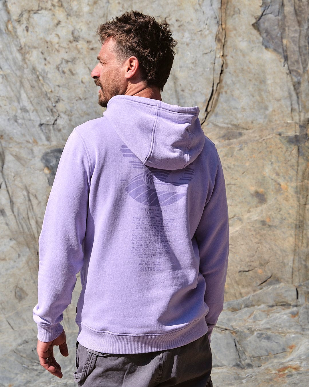 A man wearing a Saltrock - Atlantic Mens Pop Hoodie in Purple standing in front of a rock.