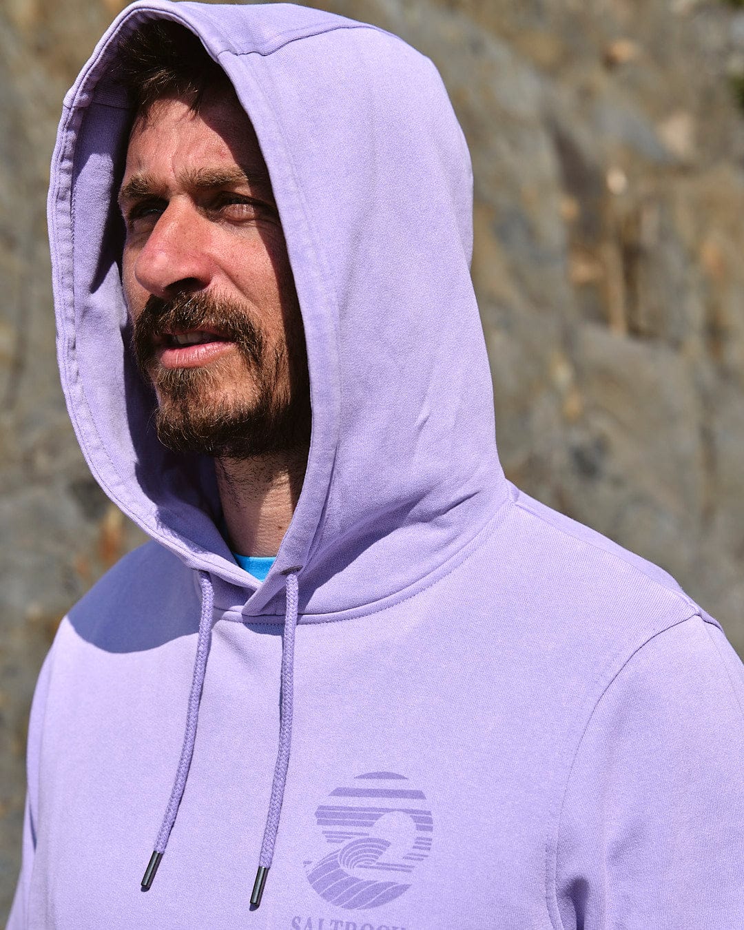A man wearing a Saltrock - Atlantic - Mens Pop Hoodie - Purple standing next to rocks.